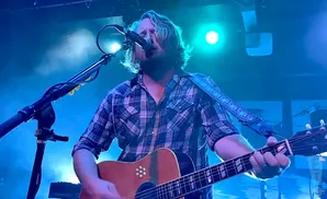 A male guitarist with long blond hair sings into a microphone while playing an acoustic guitar on a dimly lit stage.