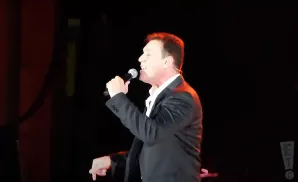 A man in a suit sings into a microphone on stage with red and black lighting.