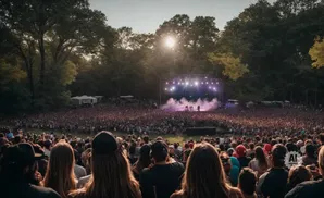 Outdoor concert with a large crowd facing a stage lit by bright lights, with trees in the background and the sun setting.
