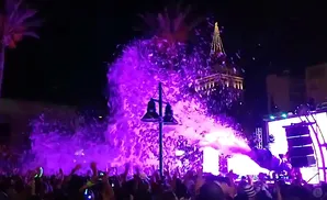 A crowd at a nighttime event watches a large plume of purple foam shoot from a stage.