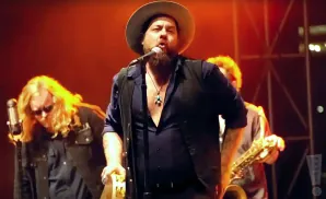 A man in a hat sings into a microphone on stage with a band.