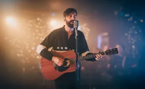 A bearded man sings into a microphone while playing an acoustic guitar on a dimly lit stage.
