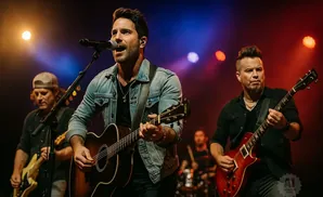 A male singer in a denim jacket performs with an acoustic guitar, backed by a band on stage.