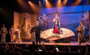 An opera performance on a stage featuring a woman in a red kimono and a man in a naval uniform, with an orchestra in the foreground.