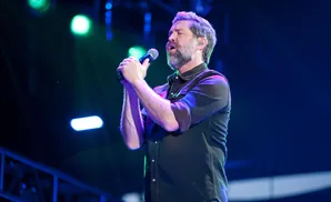 A man with a beard sings into a microphone on a stage with blue and green lights.