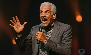 A man with gray hair holds a microphone and gestures with an open hand, speaking into it with his mouth wide open.
