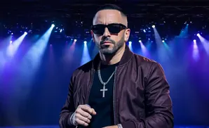 A man with sunglasses and a cross necklace stands on a stage with purple lights.