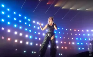 A blonde singer in a black bodysuit and chaps performs on stage with a backdrop of bright, colorful lights.