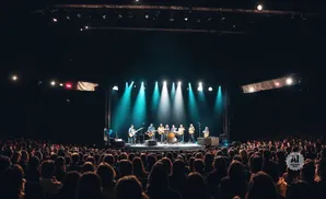 A band plays on a brightly lit stage to a large, silhouetted audience in a dark venue.