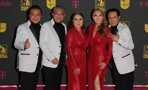 A group of four people, two women in red dresses and two men in patterned jackets, pose on a pink carpet.