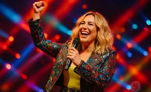 A woman with blonde hair sings into a microphone, raising her fist in the air.