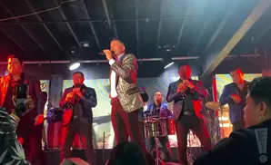 A male singer in a sequined jacket performs on stage with a band playing brass instruments and drums.