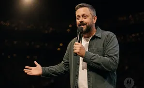 Man in denim shirt holds microphone and gestures to the left on a dark stage with blurred lights.