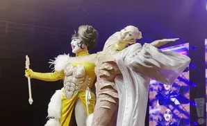 Two drag performers on stage, one in a sparkling yellow outfit with a feather boa and a staff, the other in a draped white costume.