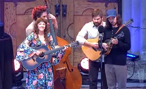 A band performs bluegrass music: a woman on guitar, a man on bass, and two men playing mandolins.