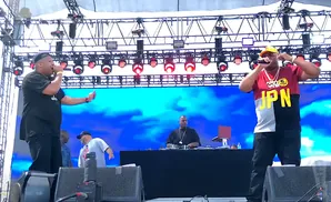 Rappers perform on stage with a DJ and a backdrop of blue clouds.