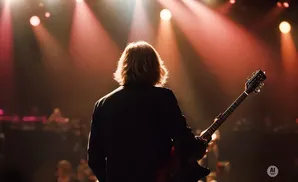 A guitarist with long blonde hair plays an electric guitar on a dimly lit stage with red spotlights.