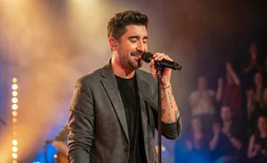 A man in a grey suit sings into a microphone on stage with warm lighting and a blurred audience.