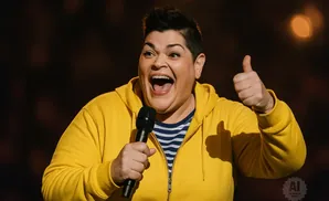 A person in a yellow hoodie smiles and gives a thumbs-up while holding a microphone.
