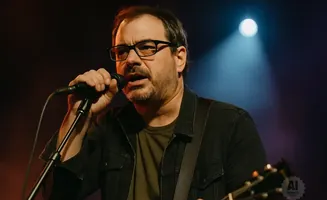 A man with glasses and a beard sings into a microphone while playing a guitar on stage.