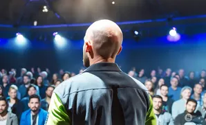 A bald man with a beard addresses a crowd from a stage in a dimly lit auditorium.