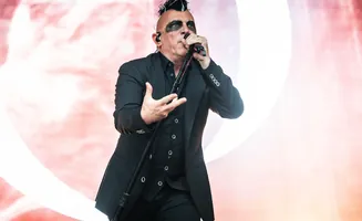 A man with a mohawk and dark eye makeup sings into a microphone on a stage, wearing a black suit.