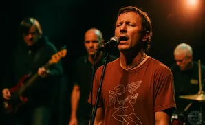 Man in a red shirt singing into a microphone with his eyes closed, band members behind him.