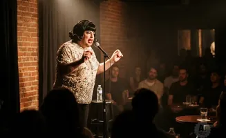 Drag performer in a sequined outfit performs on stage in a dimly lit club to an audience.