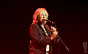 A woman with blonde hair sings into a microphone on a dimly lit stage.