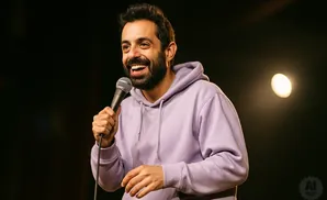 A smiling man with a beard holds a microphone and wears a light purple hoodie.