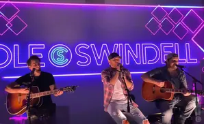 Cole Swindell performing with two other musicians on stage with a neon sign in the background.