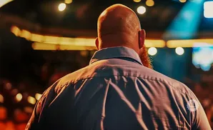 Bald man with ginger beard from behind, wearing a blue shirt under stage lights.