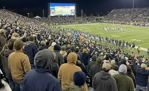Spectators watch a night football game from stadium bleachers, with a large scoreboard visible.