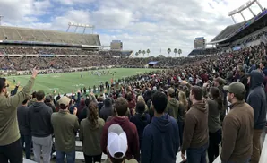 A panoramic view of a football stadium crowd with people raising their hands.