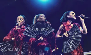 Three women in sparkly red outfits sing into microphones, holding black fans with white text.