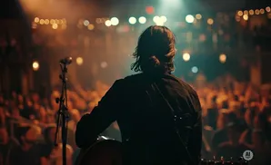A musician plays guitar on stage in front of a blurred audience with warm stage lighting.