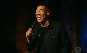 A Black man in a denim jacket laughs into a microphone on a dark stage.