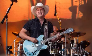 A man in a cowboy hat plays a light blue electric guitar on stage with a band.