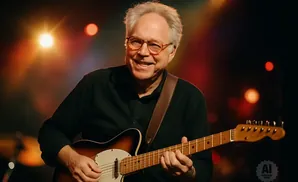 Man with glasses playing a sunburst electric guitar on a dimly lit stage with bokeh lights.