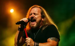 A male singer with long blonde hair and a beard sings into a microphone on a stage with orange and green lighting.