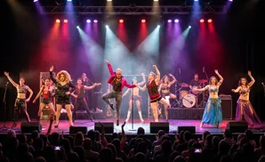 A drag queen and dancers perform on stage with a band in the background, under colorful stage lights.