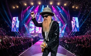 A rock guitarist with a long beard and hat performs on stage with a large crowd and colorful lights.