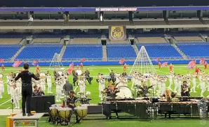 A marching band performs in a stadium, with brass instruments and percussion visible on the field.