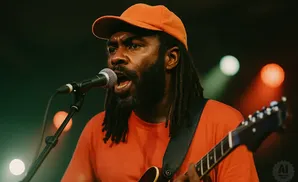A Black man with dreadlocks and an orange hat sings into a microphone while playing a guitar.