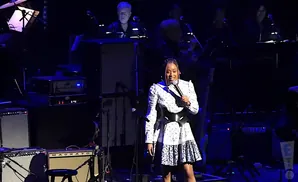 A Black woman in a floral dress sings into a microphone on a stage with musical equipment.