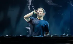 DJ Tiësto performing live on stage, singing into a microphone at a DJ booth with a large screen behind him.