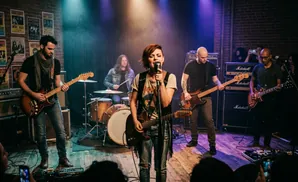 A band performs on a dimly lit stage with a singer in the foreground, guitarists and a drummer in the background.