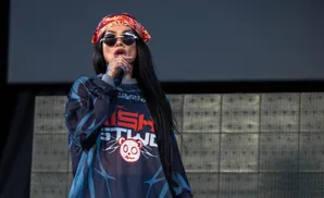 Woman wearing sunglasses and a bandana on stage, singing into a microphone.