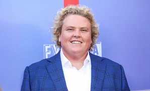 A smiling person with curly blonde hair wears a blue plaid suit jacket over a white shirt.