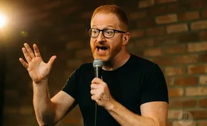 A redheaded man with glasses and a beard, holding a microphone and gesturing with his hand.